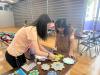 The tutor is patiently teaching the mother how to make the handicraft. 