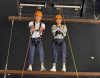 Two S3 students strike poses atop the high‑ropes beams — courage, balance and big smiles on the challenge course.