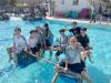 Grinning students ride their homemade bamboo-and-barrel raft in the pool, celebrating teamwork and a successful launch.