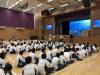 Students sit together in the school hall, listening to speakers.